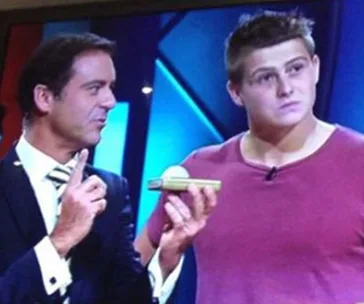 Two men stand on a TV game show set; one holds a check.