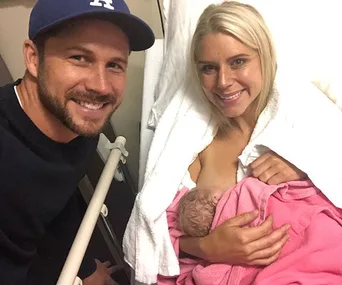 Man and woman smiling with newborn baby wrapped in pink blanket at hospital.