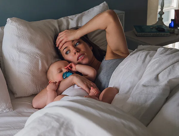 A mother lies in bed looking tired, with a baby sucking on a pacifier resting beside her.