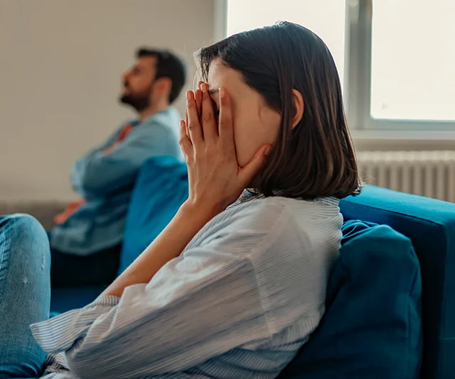 A woman sits on a sofa, covering her face with her hands, while a man sits in the background, looking away.