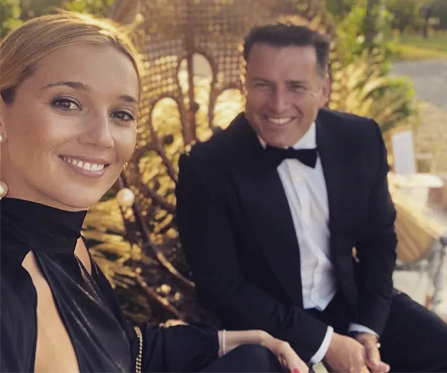 Smiling couple dressed formally; woman takes a selfie while man sits in the background.
