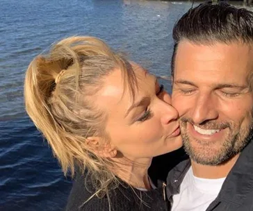 A woman kisses a smiling man on the cheek by a waterfront, both appearing happy and relaxed.