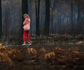Young girl in red pants and pink hoodie stands amidst charred forest, arms crossed, in aftermath of Mallacoota bushfires.