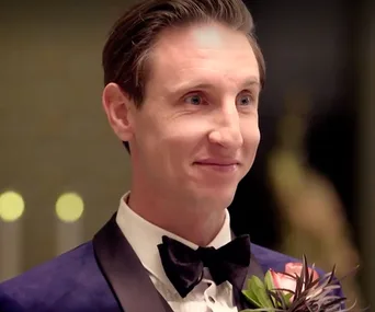 A man in a tuxedo smiles during a formal event, wearing a black bow tie and a flower boutonnière.