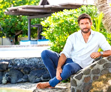 ```html
<img src="australian-survivor-all-stars-jonathan-lapaglia-return-62305" alt="Man in a white shirt sitting outdoors on a stone ledge, with lush greenery in the background." />
```