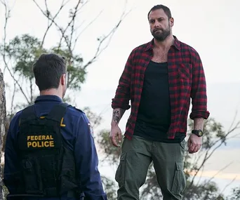 Federal officer and a man in a red plaid shirt converse outdoors in a tense scene from "Home and Away."