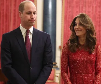 Two people standing, one in a suit and the other in a red dress, in a richly decorated room with red curtains.