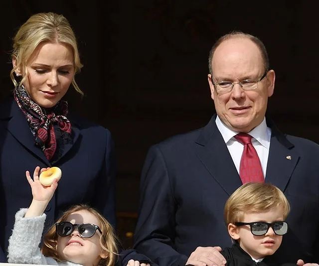 A family stands on a balcony: a woman, a man in a suit, and two children wearing sunglasses, one holding a toy.