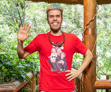 Man with multi-colored hair, wearing a red t-shirt with a printed image, smiling and waving in an outdoor setting.