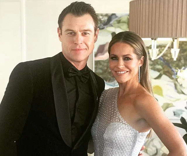 A couple dressed elegantly, with the man in a black tuxedo and the woman in a silver one-shoulder dress, smiling indoors.