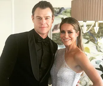 A couple dressed elegantly, with the man in a black tuxedo and the woman in a silver one-shoulder dress, smiling indoors.