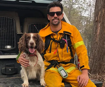 Firefighter in yellow gear with search and rescue dog, sitting on a vehicle, trees in the background.