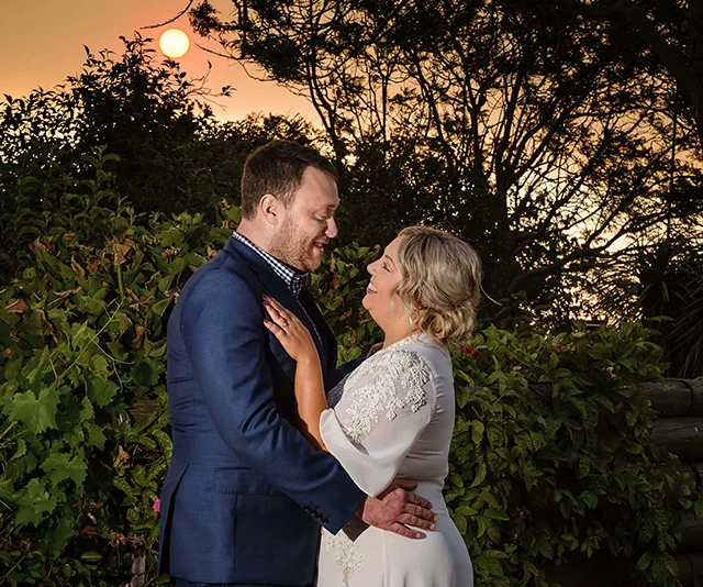 A couple embraces at sunset, surrounded by greenery with a glowing sky in the background.