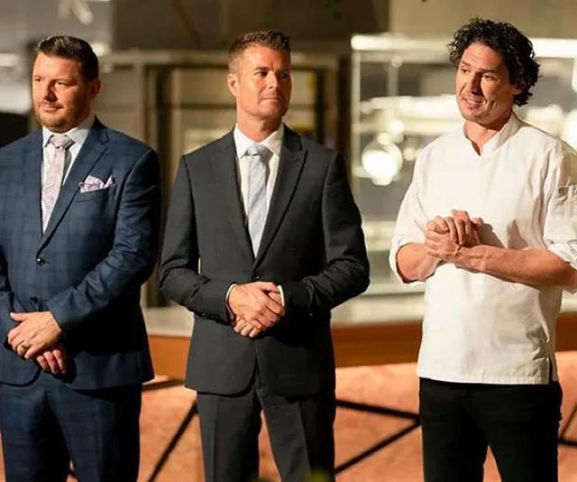 Three judges from a cooking show stand together; two in suits, one in a chef's jacket.