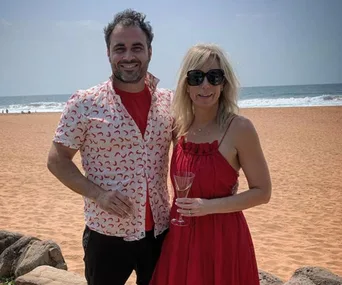 Man and woman in red outfits smiling on a beach; woman holding a glass.