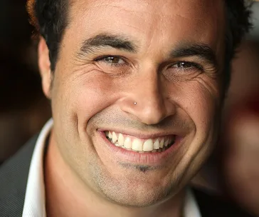 Smiling person with a nose stud and facial hair, wearing a dark jacket and white shirt.