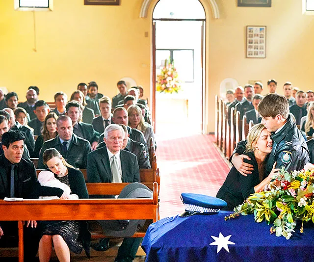 Funeral scene with mourners seated, a casket draped in an Australian flag, and two people embracing at the front.