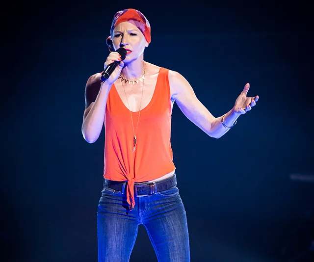Singer in orange top and bandana performs on stage holding a microphone.