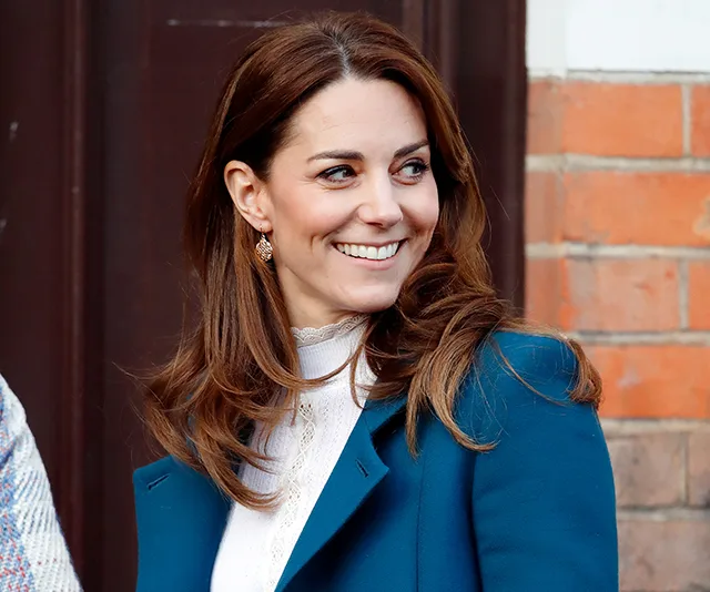 A woman in a teal coat smiling, standing outside against a brick and wooden backdrop.