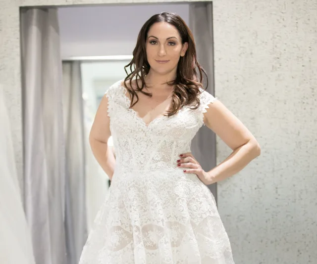 A woman in a bridal gown stands confidently in a room with light-colored walls and curtains.
