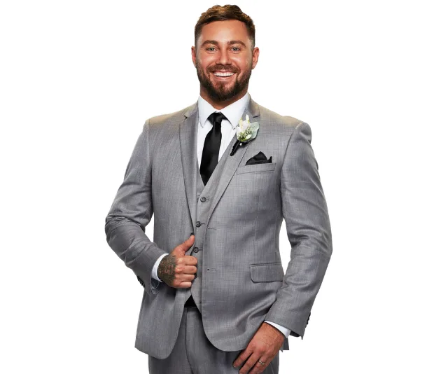 Man in a gray suit with a flower boutonniere, smiling confidently against a white background.