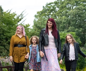 Four people walking outdoors, including two adults and two children, with trees in the background.