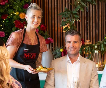 Kerry serving food at a table with a formally-dressed man, decorated with flowers and wooden paneling in the background.