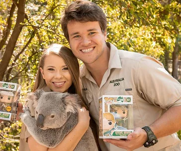 Two people smiling, holding Steve Irwin Funko Pop figures, with a koala, wearing Australia Zoo shirts.