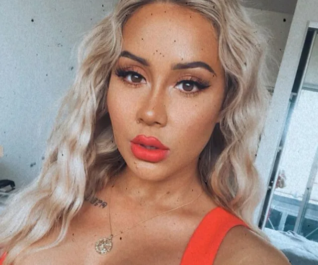 Blonde woman with wavy hair and bright red lipstick, wearing a red top, poses indoors.