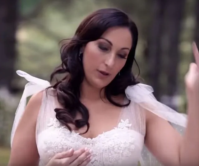 A bride in a white gown with flowing sleeves appears to be adjusting her dress outdoors in a wooded area.