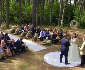 Outdoor wedding ceremony in a forest setting with guests seated on wooden benches, a couple standing at the altar.