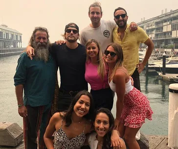 Group of friends pose together by the waterfront, with a marina and buildings in the background.