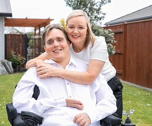 Man in a wheelchair smiling, embraced by a woman, in a garden setting.