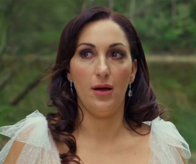 A woman with long brown hair and earrings looks surprised, standing outdoors in a white dress.