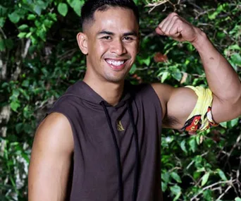 Man smiling, flexing bicep, with a yellow armband, in front of greenery.