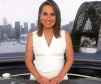 A smiling person in white attire stands at a news desk with a cityscape and bridge in the background.