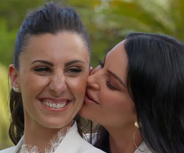 Two women smiling, one kissing the other's cheek, wearing white outfits, outdoors with greenery in background.