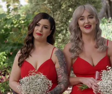 Two women in red dresses holding flowers, one with a tattoo, smiling outdoors.
