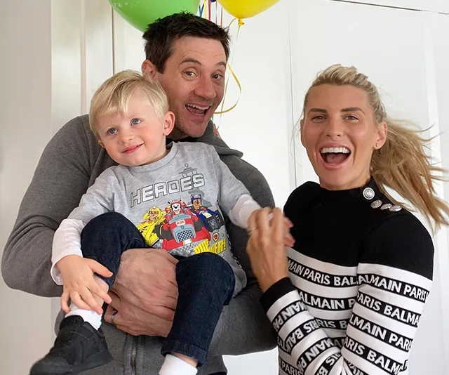 A family smiling and holding a child with balloons in the background.