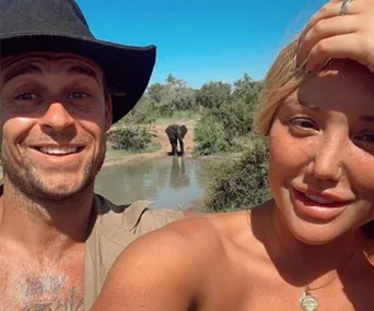 Selfie of a man and woman outdoors with an elephant in the background near a water hole.