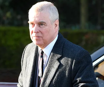A man in a suit and tie outside, wearing a dark coat, in bright daylight with trees in the background.