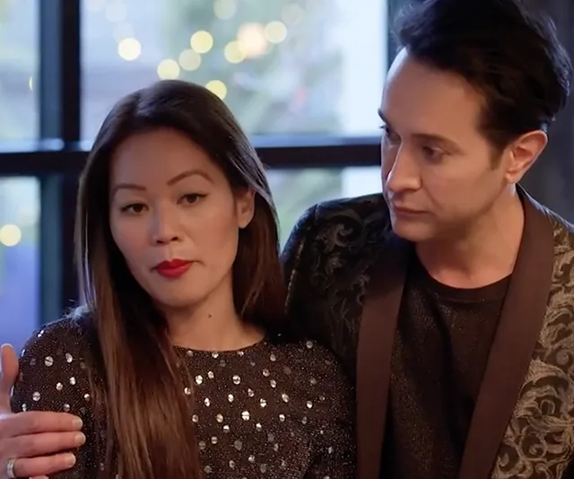A woman in a sparkly top is talking while a man in a dark jacket looks at her, with blurred lights in the background.