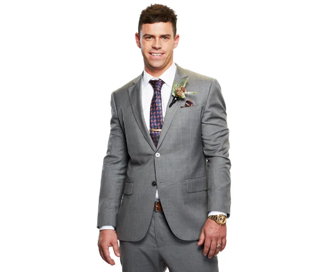 Man in a gray suit with a floral boutonniere smiles confidently, wearing a watch and a patterned tie.
