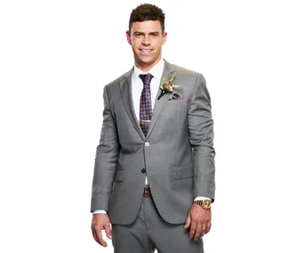 Man in a gray suit with a floral boutonniere smiles confidently, wearing a watch and a patterned tie.
