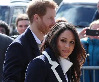 "Two well-dressed individuals outdoors, surrounded by a crowd, one with dark hair wearing a navy coat."