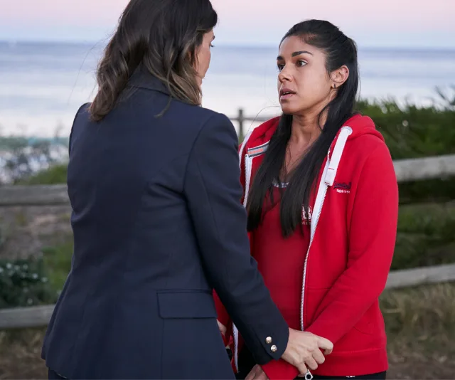 Two women stand facing each other near a seaside fence, one holding the other's hands, in a tense conversation.