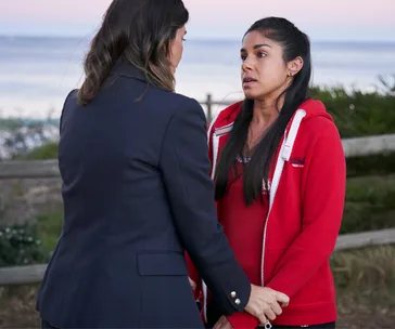 Two women stand facing each other near a seaside fence, one holding the other's hands, in a tense conversation.