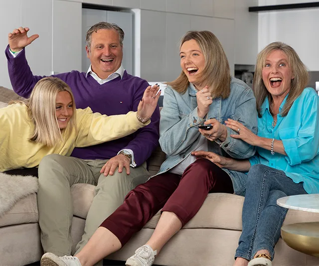 A family of four sitting on a couch, laughing and holding a TV remote, appearing to enjoy a show together.