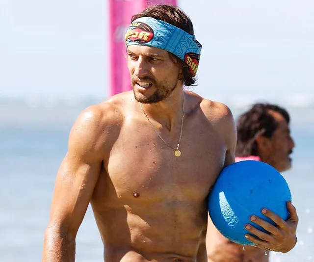 Man with a 'Survivor' headband holding a blue ball on a beach.