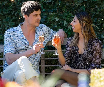 Two people sitting outdoors on a bench, toasting with glasses of rose wine.